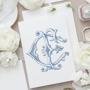 May include: A white rectangular card featuring a blue ink illustration of an ornate, intertwined monogram letter design. The card is surrounded by white ranunculus flowers, a small velvet ring box with a diamond ring, and a gold shield emblem.