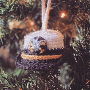 May include: A crocheted ornament shaped like a hat with a white brim, a gold band, and a blue and white center. The ornament is hanging on a green Christmas tree.