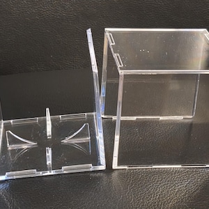 May include: Two clear acrylic display boxes, one assembled and one disassembled. The assembled box is a cube with a rectangular base. The disassembled box has a base with support structures and side panels. The boxes are designed to showcase items.