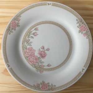 Peut inclure: Une assiette plate en céramique blanche avec un bord doré et un motif floral composé de roses roses et de feuilles vertes.