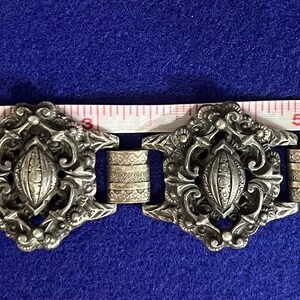 Può includere: Un braccialetto d'argento con un design decorativo che presenta intricati motivi floreali e una forma ovale centrale. Il braccialetto è lungo circa 18 cm.