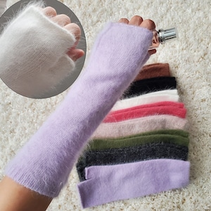 May include: A stack of eight pairs of fuzzy fingerless gloves in various colors, including white, lavender, pink, red, green, black, and brown. The gloves are made of a soft, fluffy material.