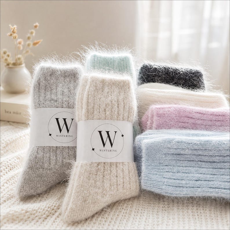 May include: A collection of fuzzy, ribbed knit socks in various colours including grey, beige, light blue, pink, white, and dark grey. Each pair features a white paper band with a black circular logo containing the letter W and the text WINTERING.