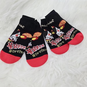 May include: Black ankle socks with red toes and a graphic of a cartoon chicken with flames. The socks have text in a foreign language. The brand name "Samyang" is printed on the top of the socks.