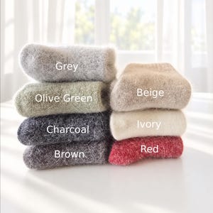 May include: A stack of six fuzzy, textured socks in various colors, labeled with text: Grey, Olive Green, Charcoal, Brown, Beige, Ivory, and Red. The socks have a soft, brushed appearance and are arranged in two columns on a white surface.