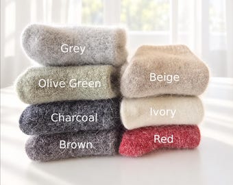 May include: A stack of six fuzzy, textured socks in various colors, labeled with text: Grey, Olive Green, Charcoal, Brown, Beige, Ivory, and Red. The socks have a soft, brushed appearance and are arranged in two columns on a white surface.