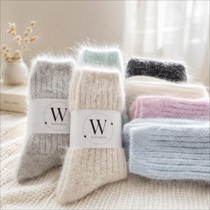 May include: A collection of fuzzy, ribbed knit socks in various colours including grey, beige, light blue, pink, white, and dark grey. Each pair features a white paper band with a black circular logo containing the letter W and the text WINTERING.