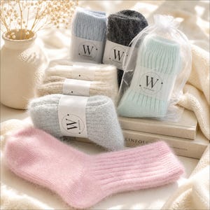 May include: A collection of fuzzy, ribbed knit socks in various pastel and neutral colours including pink, light blue, grey, cream, and mint green. Each pair features a white paper band with the letter W and the text WINTERING printed on it.