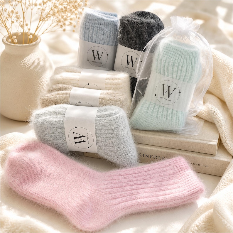 May include: A collection of fuzzy, ribbed knit socks in various pastel and neutral colours including pink, light blue, grey, cream, and mint green. Each pair features a white paper band with the letter W and the text WINTERING printed on it.