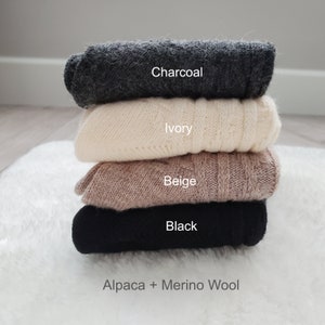 Pode incluir: Uma pilha de quatro su&eacute;teres de alpaca e l&atilde; merino em diferentes cores: carv&atilde;o, marfim, bege e preto. Os su&eacute;teres est&atilde;o dobrados e empilhados um em cima do outro. O texto "Alpaca + Merino Wool" est&aacute; impresso abaixo da pilha de su&eacute;teres.