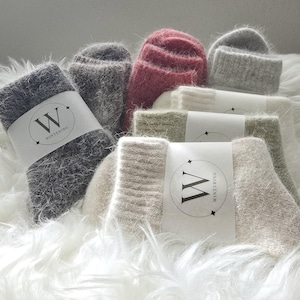 May include: A collection of fuzzy, soft-textured socks in various colors including dark gray, rose pink, light gray, cream, and beige. Each pair features a white paper band with the letter W and the text WINTERING printed in black.