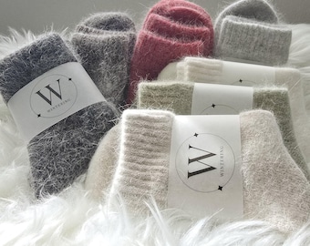 Puede incluir: Una colección de calcetines difusos de textura suave en varios colores, incluyendo gris oscuro, rosa, gris claro, crema y beige. Cada par cuenta con una banda de papel blanca con la letra W y el texto WINTERING impreso en negro.