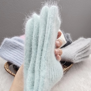 May include: A pair of light mint green fuzzy knit socks with a ribbed texture. The socks are held up vertically, showcasing the soft, long-fibre material. Other folded pairs of socks in shades of light blue, pink, white, and grey are visible.