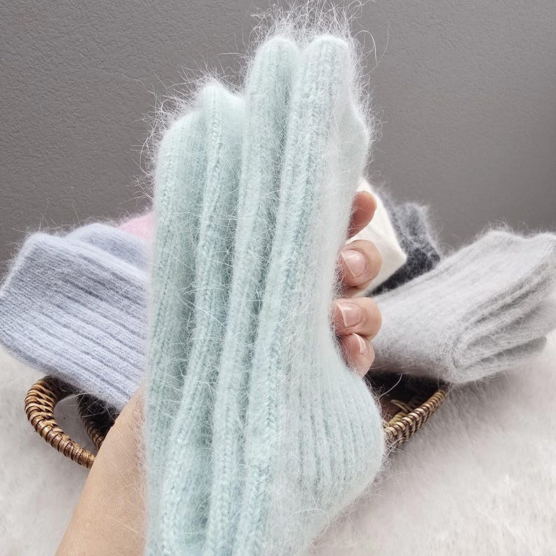 May include: A pair of light mint green fuzzy knit socks with a ribbed texture. The socks are held up vertically, showcasing the soft, long-fibre material. Other folded pairs of socks in shades of light blue, pink, white, and grey are visible.
