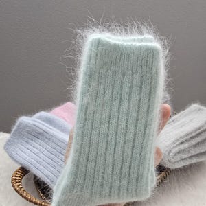 May include: A pair of fuzzy, ribbed knit socks in a soft mint green color. The material has a long, soft pile texture. Other pairs of socks in light blue, pale pink, and heather gray are visible in the background, all featuring the same ribbed knit pattern and fuzzy texture.