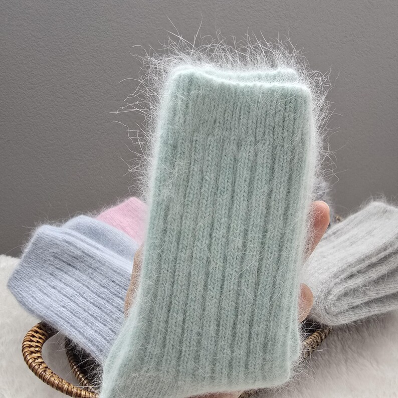May include: A pair of fuzzy, ribbed knit socks in a soft mint green color. The material has a long, soft pile texture. Other pairs of socks in light blue, pale pink, and heather gray are visible in the background, all featuring the same ribbed knit pattern and fuzzy texture.