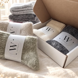 May include: A collection of fuzzy, knit socks in shades of cream, sage green, charcoal, and light gray. Each pair is wrapped in a white paper band featuring a circular logo with the letter W and the text WINTERING. Some socks are neatly folded inside a white cardboard box, while others are stacked outside.