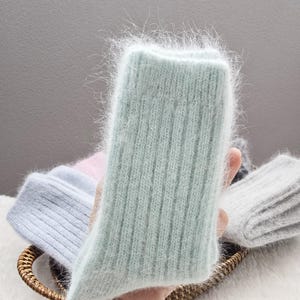 May include: A pair of fuzzy, light mint green ribbed socks with a soft, brushed texture. The socks feature a vertical knit pattern and a thick, plush appearance. Other folded socks in shades of light blue, pink, and grey are visible nearby.