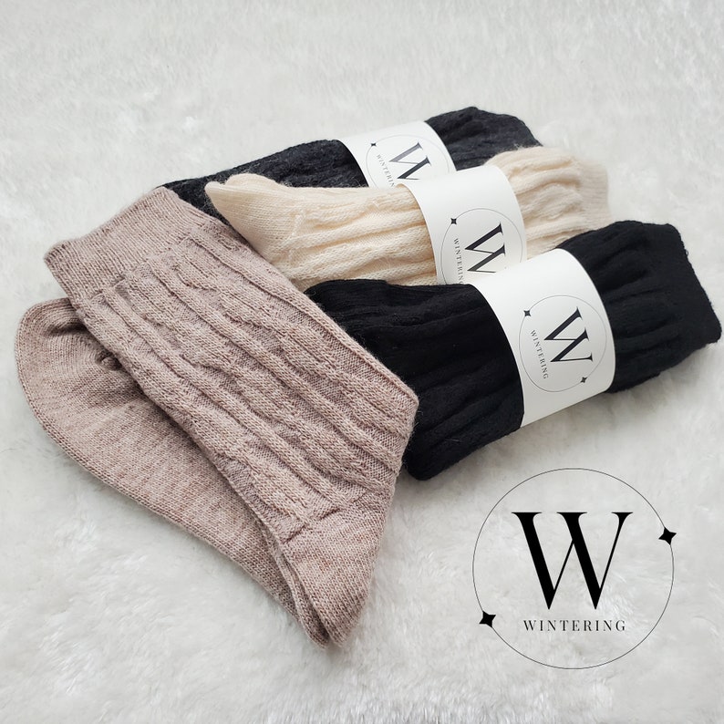Pode incluir: Tr&ecirc;s pares de meias em bege, preto e branco com um padr&atilde;o de tric&ocirc; tran&ccedil;ado. Cada par tem uma etiqueta branca com um "W" preto e a palavra "WINTERING" nela.