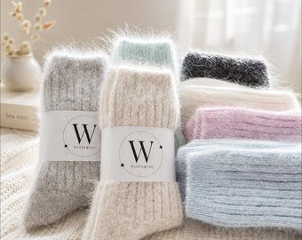 May include: A collection of fuzzy, ribbed knit socks in various colors including gray, beige, mint green, charcoal, white, pink, and light blue. Each pair features a white paper band with the letter W and the text WINTERING printed on it.