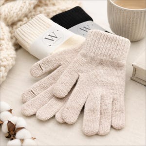 May include: A pair of beige knit gloves with a ribbed cuff, featuring a subtle speckled texture. Behind them, two additional pairs of gloves are visible, one in cream and one in black, each wrapped in a white paper band with the letter W.