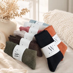 May include: A stack of folded socks in various colors including black, olive green, dark brown, light gray, dusty rose, white, and burnt orange. Each pair is secured with a white paper band featuring the letter W and the text WINTERING.