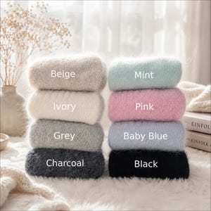 May include: Two stacks of folded fuzzy sweaters in eight colors. The left stack features beige, ivory, grey, and charcoal. The right stack features mint, pink, baby blue, and black. Each sweater is labeled with its corresponding color name.