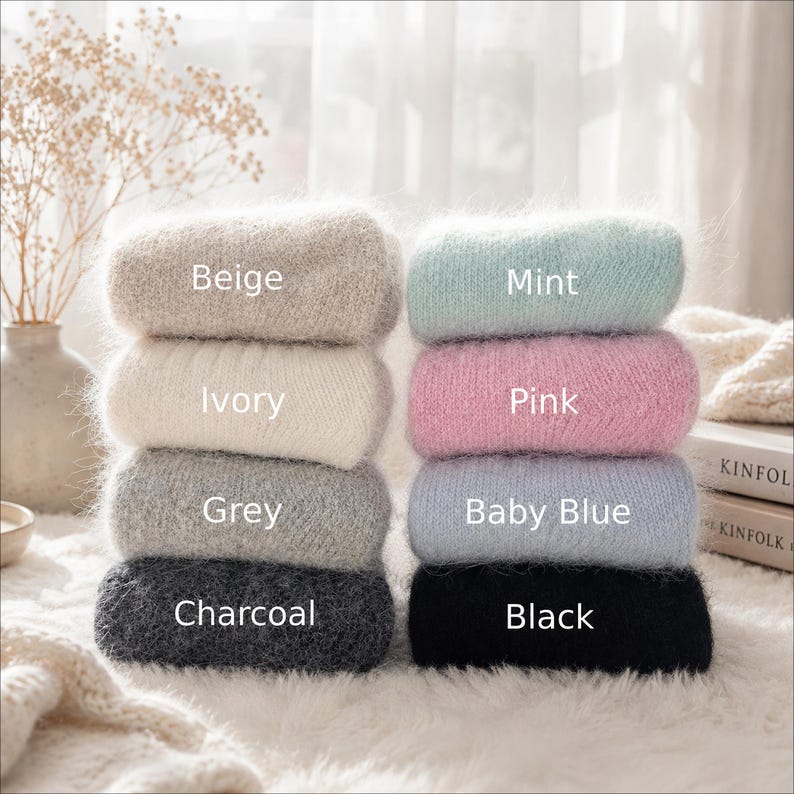 May include: Two stacks of folded fuzzy sweaters in eight colors. The left stack features beige, ivory, grey, and charcoal. The right stack features mint, pink, baby blue, and black. Each sweater is labeled with its corresponding color name.