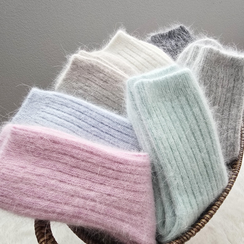 May include: A collection of fuzzy, ribbed knit socks in various pastel and neutral colours, including soft pink, light blue, mint green, beige, white, and grey. The socks are arranged in a woven basket, showcasing their soft, textured material.