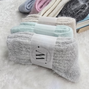 May include: A collection of fuzzy, ribbed knit socks in various colours including light blue, pink, cream, black, beige, mint green, and grey. Each pair is folded and secured with a white paper band featuring a circular logo with the letter W and the text WINTERING.