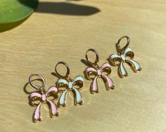 May include: Four pairs of gold-colored hoop earrings with bow-shaped pendants. The bows are white and pink with gold outlines. The earrings are displayed on a light wooden surface, with a green leaf in the upper left corner.