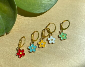 May include: Five gold-colored hoop earrings, each with a different colored flower charm: red, blue, yellow, white, and green. The earrings are arranged in a row on a light-colored wooden surface. The earrings are a fun and colorful accessory.