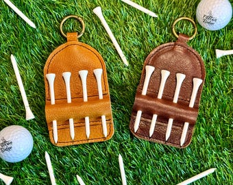 May include: Two leather golf tee holders, one tan and one brown, each holding white golf tees. Golf balls and loose tees are scattered on a green surface. The holders have a metal ring for attachment.