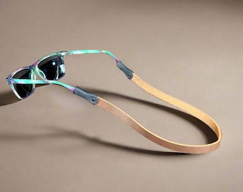 Peut inclure: Lunettes de soleil avec une lanière en cuir marron clair. Les lunettes ont une monture colorée à motifs avec des verres foncés. La lanière est fixée à la monture par des connecteurs gris. Le fond est marron uni.
