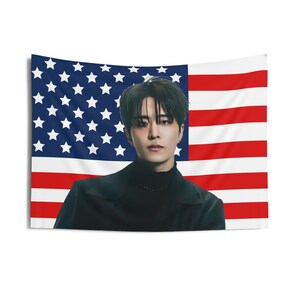 Könnte beinhalten: Ein Mann mit schwarzen Haaren steht vor einer amerikanischen Flagge. Er trägt einen schwarzen Rollkragenpullover und eine schwarze Jacke.