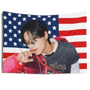 May include: A person with black hair wearing a gray shirt with the text "The Ones" on it, a pink jacket, and a blue and white striped face mask. The person is standing in front of an American flag.