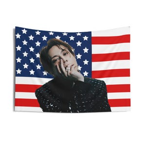 Könnte beinhalten: Ein Mann mit braunen Haaren trägt eine schwarze Jacke mit Muster. Er posiert vor einer amerikanischen Flagge.