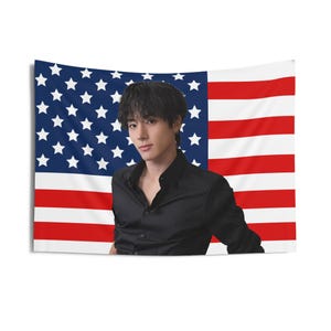 May include: A man with dark hair is standing in front of an American flag. He is wearing a black button-down shirt.