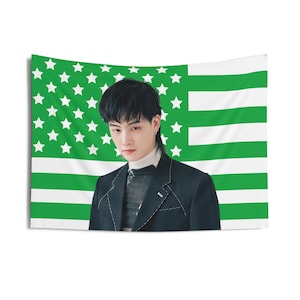 Op de afbeelding: Een man met zwart haar, gekleed in een zwart blazer, staat voor een Amerikaanse vlag met groene en witte strepen. De vlag heeft witte sterren op een groene achtergrond.