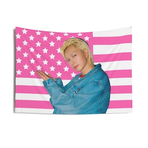 May include: A pink and white American flag tapestry with a person in a denim jacket. The flag design features pink stripes and white stars. The person is wearing a red shirt under the jacket.