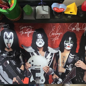 May include: A rectangular desk mat featuring a color photograph of the rock band KISS in full makeup and stage costumes against a red background. The image includes four band members with their printed signatures above them on a dark surface.