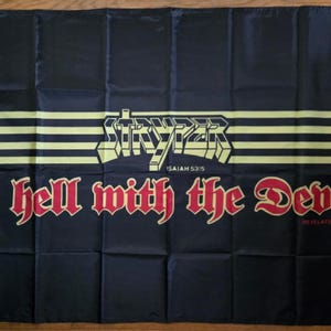 May include: A rectangular black fabric flag featuring the yellow text STRYPER above the phrase Isaiah 53:5. Below this, the red text to hell with the Devil is printed in a stylized font, followed by the small red text Revelation 20:10.