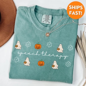 May include: A teal Comfort Colors t-shirt featuring a Halloween-themed design. The shirt has a graphic of ghosts, pumpkins, and flowers, with the words "speech therapy" in a script font. A silver necklace with a pendant is also visible.