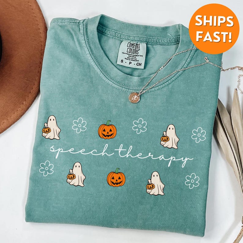 May include: A teal Comfort Colors t-shirt featuring a Halloween-themed design. The shirt has a graphic of ghosts, pumpkins, and flowers, with the words "speech therapy" in a script font. A silver necklace with a pendant is also visible.