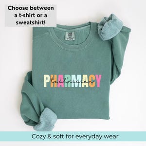 May include: A teal green sweatshirt with the text "PHARMACY technician" in rainbow colours. The sweatshirt is folded and displayed on a white background.
