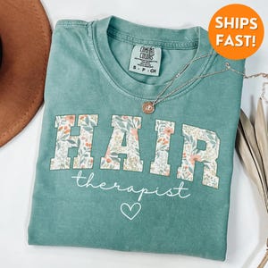 May include: A teal green t-shirt with a floral print design that says "HAIR therapist" in white text with a small heart below the text.