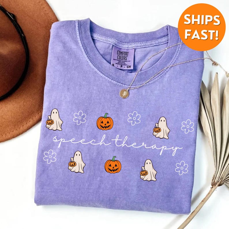 May include: A lavender t-shirt with a crew neck featuring a design of cartoon ghosts, pumpkins, and flowers. The words "speech therapy" are written in a cursive font. A gold necklace is draped across the shirt.