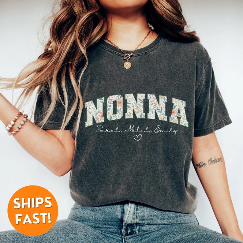 May include: A dark gray t-shirt with a floral print design. The word "NONNA" is printed in white with a black outline. The names "Sarah, Mitch, Emily" are printed in white below the word "NONNA".