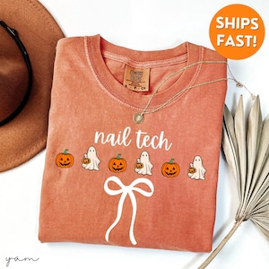 May include: A coral-coloured sweatshirt featuring the text "nail tech" in white. The design includes orange pumpkins and white ghost figures. A white bow graphic is below the text. A gold necklace is draped on the shirt.