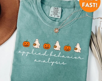 May include: Light green t-shirt with a graphic of pumpkins and ghosts. The text "applied behavior analysis" is printed below the graphic. A silver necklace with a pendant is on the shirt.